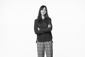A black and white photo of Carilyn Brandt, standing against a white background with her arms crossed and smiling at the camera.