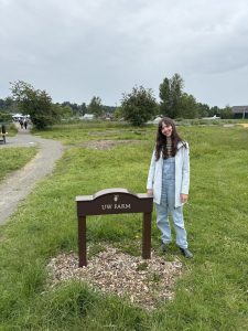 Carilyn Brandt stands on the grass next to the UW farm sign, with a gravel path and trees in the background.