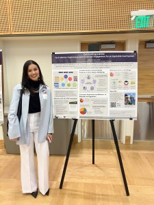 Stephanie stands next to her poster, posing for the camera and smiling. Her poster is large and mounted on an easel behind her. 