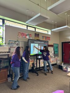 3 students in purple shirts stand at the front of a classroom. One of them is pointing at a map being displayed on a TV projector. 