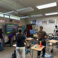 Students stand next to desks in a classroom setting. There are 3 instructors displaying a lesson on a TV at the front of the room.