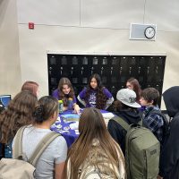Students and teachers stand around a tall round table looking at an assortment of stickers and brochures.