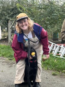 Kaija Koenigberg bends towards the camera with a smile on their face, petting a  large black dog sitting between their legs. They are outside with trees and grass in the background. 