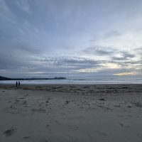 An image of a sandy beach at sunset, with a cloudy ski and two people in the background walking.