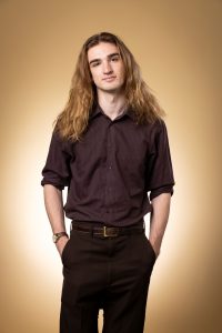 Ethan stands against a gold background with his hands in his pockets, looking at the camera. He wears a brown shirt and black pants. 