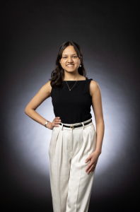 Sonali stands against a black background with her hand on her hip, smiling at the camera. She wears a black shirt and white pants. 