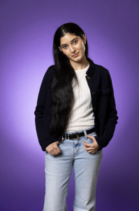 Sarah stands against a bright purple background, posing and looking at the camera. She wears a white shirt, black jacket, and jeans. 