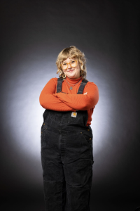 Kaija stands against a black background, arms crossed and smiling at the camera. They wear an orange shirt and black overalls. 