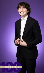 Cillian stands against a bright purple background and smiles at the camera. He wears a black suit with a white undershirt. 