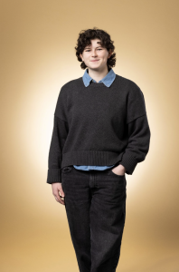 Mathilda stands against a gold background with one hand in her pocket, looking at the camera. She wears a grey sweater and black pants. 
