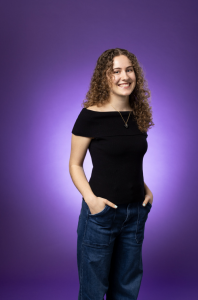 Lorin stands against a bright purple background and smiles at the camera. She wears a black shirt and dark jeans. 