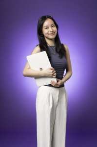 Farrah stands against a bright purple background and smiles at the camera. She holds a laptop in one hand. 
