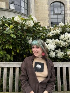 Kalina sits on a bench outside in front of a green bush with white flowers. She smiles and wears a brown shirt. 