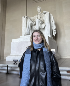 Seiler stands smiling at the camera, wearing a black leather jacket and blue scarf. In the background is the Lincoln memorial statue. 