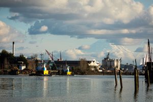A photograph of the Duwamish River, with water in the foreground and mount rainier in the background. There is industry along the river and a couple of tug boats in the water.